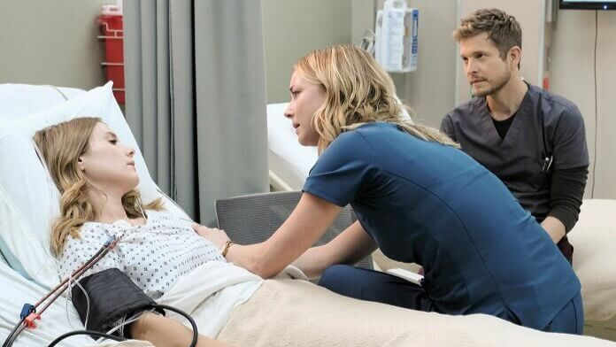 In this scene from The Resident, a blonde female nurse leans over a patient's hospital bed to offer comfort. A male doctor watches closely from behind, creating a serious and emotional medical atmosphere.
