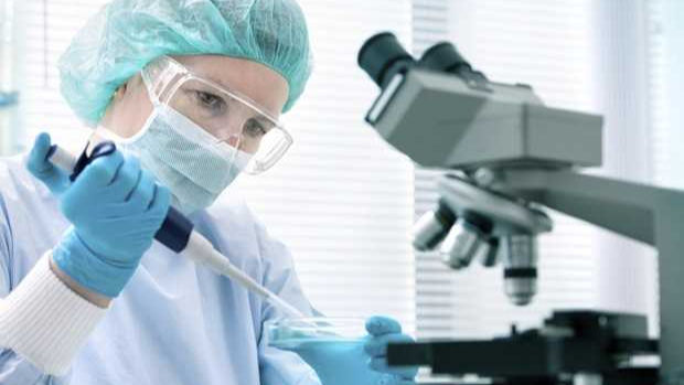 A scientist in protective gear, including a hairnet and goggles, is using a pipette to work with a petri dish. A microscope is visible in the background of the lab.