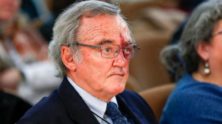 Un primer plano muestra a un hombre mayor con gafas y una marca de nacimiento rojiza en el rostro, vistiendo traje oscuro y corbata, mientras escucha atentamente en lo que parece ser una conferencia.