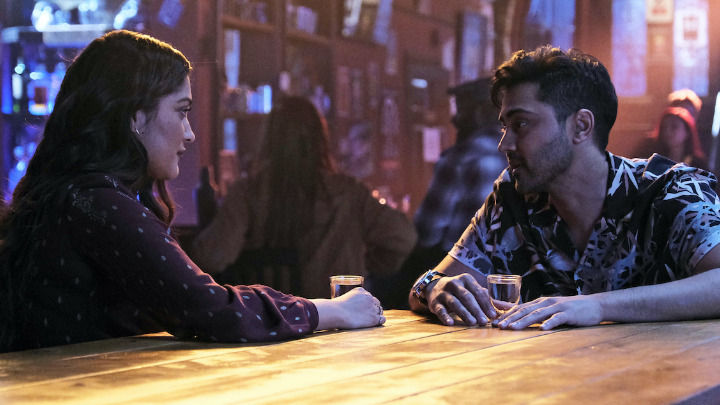 In a dimly lit bar, a young man and woman sit across from each other at a wooden table with small glasses. They are engaged in a close, personal conversation with focused and thoughtful expressions.