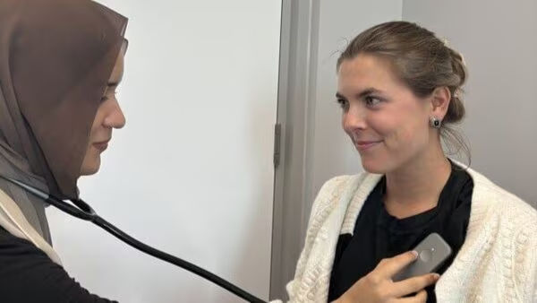 This photo shows a doctor in a hijab using a stethoscope on a smiling patient. The patient holds what appears to be an AliveCor portable ECG monitor, used for checking heart rhythm.