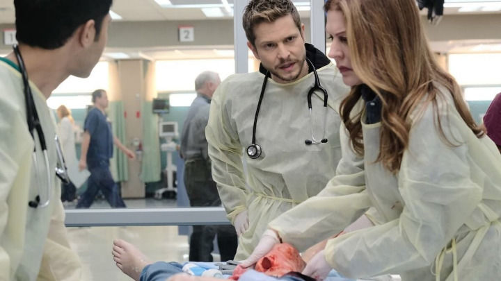 En un hospital concurrido, tres profesionales médicos con batas protectoras amarillas y estetoscopios atienden urgentemente a un paciente con una herida abdominal grave, mostrando una concentración intensa durante esta situación de emergencia crítica.