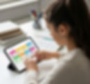 A young girl with dark hair taps a tablet displaying a colorful electronic planner. She is seated at a white desk with books and pens nearby, indicating study.