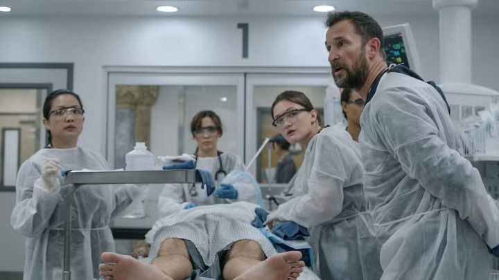 Three medical professionals, including a bearded doctor and two female nurses, are urgently treating a young male patient on a hospital bed. Their intense, focused expressions reflect a high-stakes emergency situation.