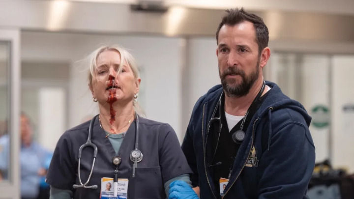 A bearded doctor wearing a navy hoodie supportively holds the arm of a blonde nurse leader who has blood running down her face. Their shocked expressions highlight a violent or traumatic incident within the hospital.