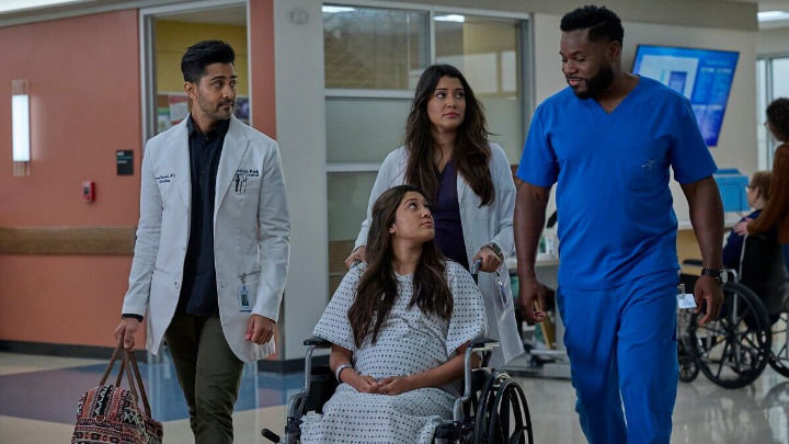 En un pasillo de hospital iluminado, tres profesionales médicos acompañan a una paciente embarazada en silla de ruedas. Un doctor lleva un bolso estampado mientras el grupo camina unido, manteniendo una conversación que parece tranquila y de apoyo.