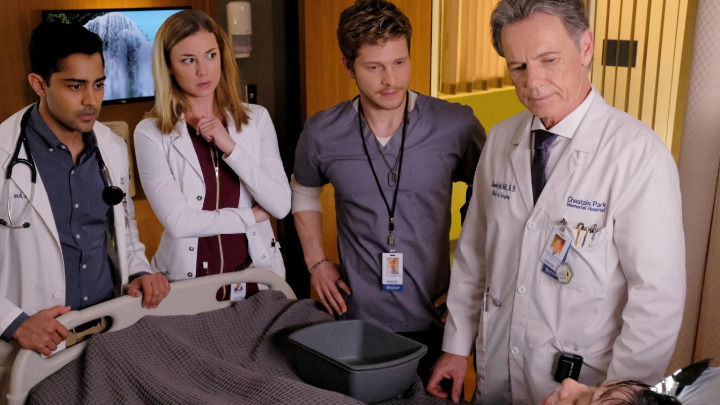 Cuatro profesionales médicos, incluyendo doctores con batas blancas y uniformes quirúrgicos, rodean una cama en el Chastain Park Memorial Hospital, observando con preocupación a un paciente que recibe atención clínica intensiva en la habitación.