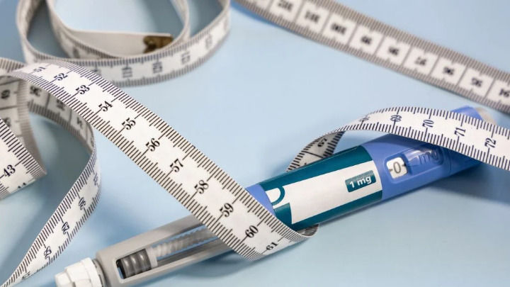 A blue medication pen labeled "1 mg" lies on a light surface, entwined with a white measuring tape. The image symbolizes medical treatment related to weight management and clinical dosage monitoring.