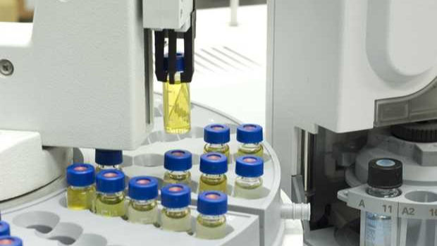 A detailed view of a laboratory machine, with a robotic arm manipulating a glass test tube containing a yellow liquid. There are more test tubes in a tray.