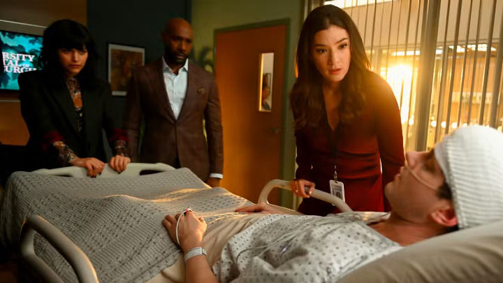 This scene shows Dr. Watson and his colleagues watching with serious faces as a patient with a bandaged head rests in a hospital bed under a warm side light in a clinic.