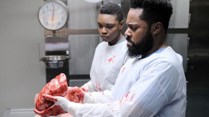 In this striking image from The Resident, Dr. AJ Austin and another medical professional in white surgical gowns hold a large, bloody organ. They look on with deep concentration and concern during a morgue procedure.
