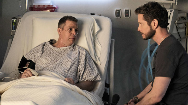 In a brightly lit clinical room, a male medical professional in a dark shirt sits beside a patient’s bed. They are engaged in a serious and personal conversation, with the patient looking at him attentively.