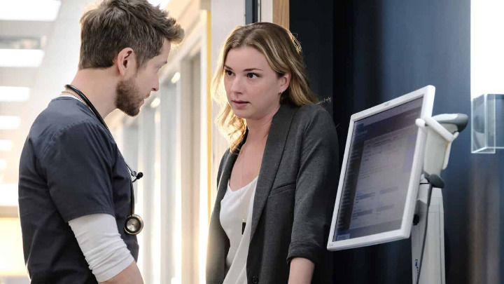 In a hospital hallway, Conrad Hawkins and Nic Nevin from The Resident share a serious conversation. They stand near a computer monitor, their focused expressions conveying the weight of a professional medical discussion.