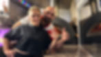 Man and woman smiling in front of wood-fired pizza ovens.