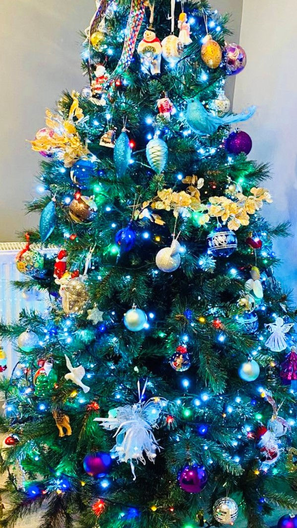 A Christmas tree, decorated in lots colourful decorations and many coloured lights. The grey-green walls are just visible in the background.