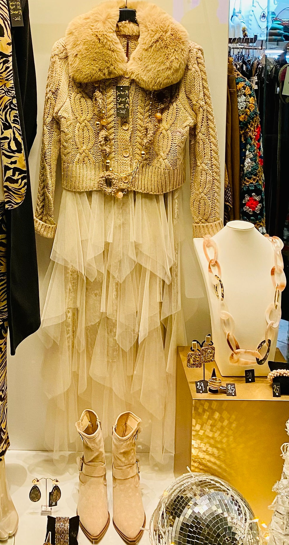 A window dressed with an outfit of a natural, cable-knit jumper with a fur collar, a netting skirt and suede boots. A disco ball in the corner casts multiple bright lights across the display..