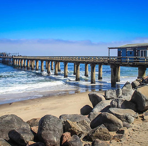 El muelle se extiende hacia el océano con edificios, cielo azul sobre la playa de arena.