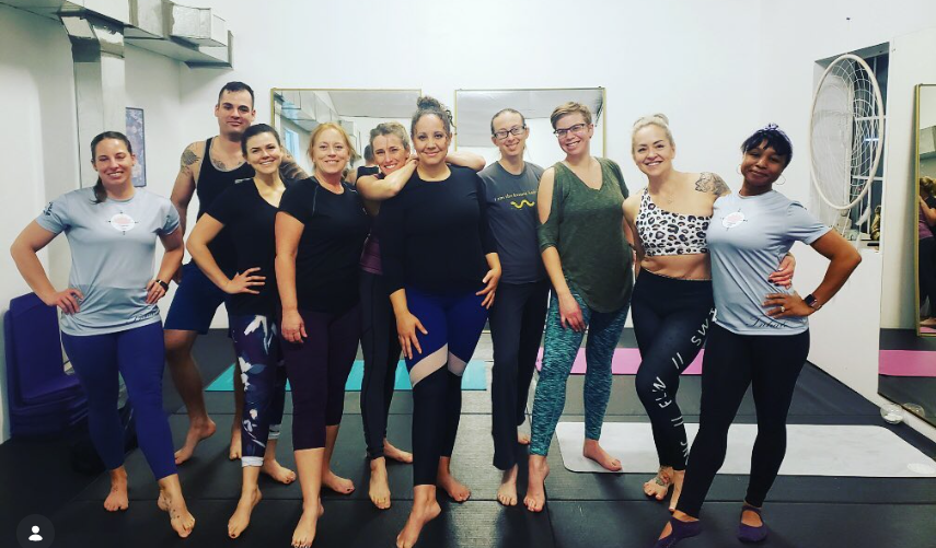 Group of eight smiling adults posing barefoot in a yoga studio with mats, mirrors, and a large fan.