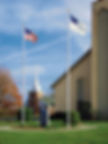 Two flagpoles outside a church, one flying the American flag and the other flying the Christian flag, with cleats, ropes, and ball toppers.