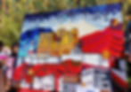 A vibrant collage artwork made from waste cloth scrap depicts a colorful cityscape with red roofs and textured buildings, set against a backdrop of dynamic mountains and a dramatic sky.