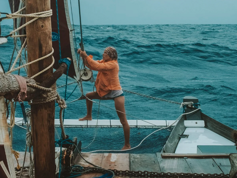 One of the three women is standing in the jib net, one leg braced against the mast, both arms holding the sail line—presumably while repairing the sail, with the blue Atlantic stretching out behind her.