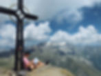 Hiker leaning against the summit cross, with a sweeping mountain panorama in the background, carrying hiking poles and writing in the summit book – an important moment in tour documentation and hiking safety.