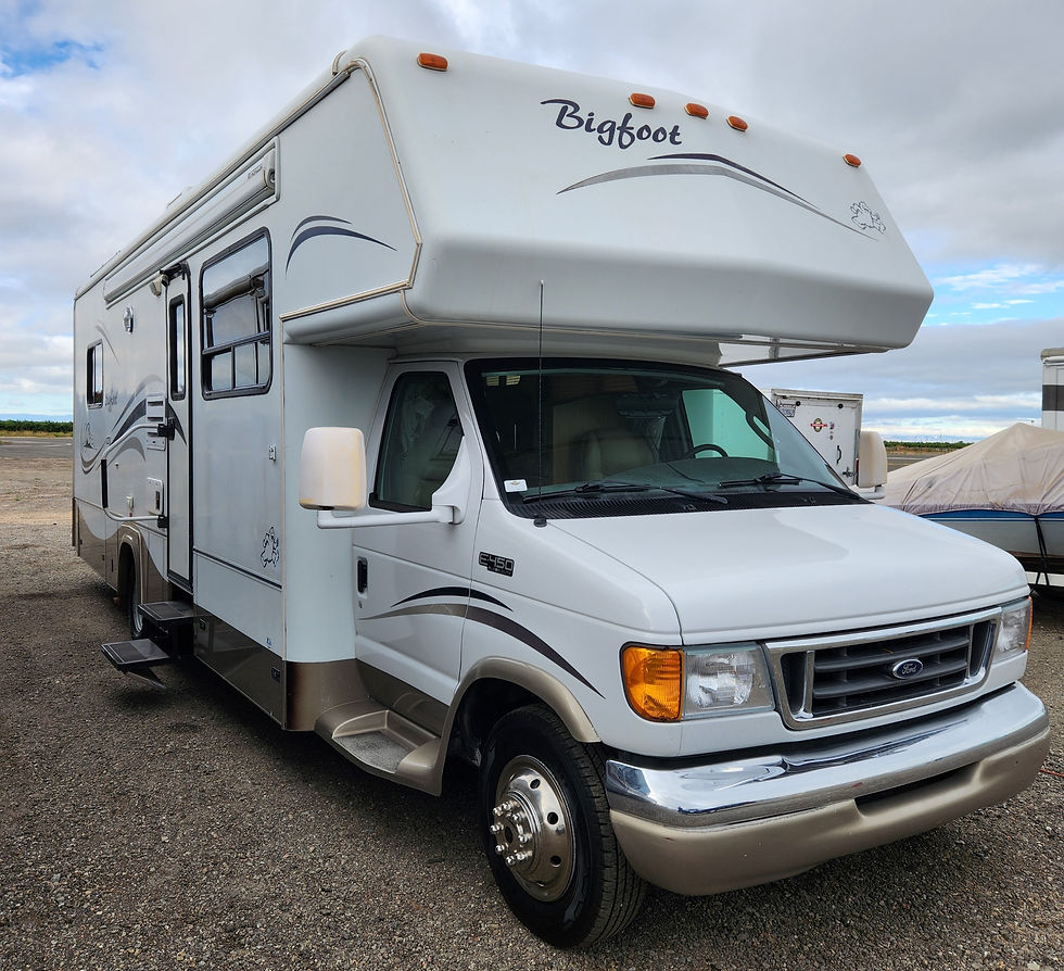White motorhome labeled "Bigfoot" parked on gravel, overcast sky. Features blue-gray swirls on sides, steps extended, serene setting.
