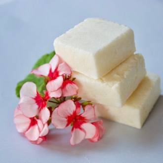 Three white soap bars stacked beside pink and red flowers on a light background, creating a fresh and soothing atmosphere.