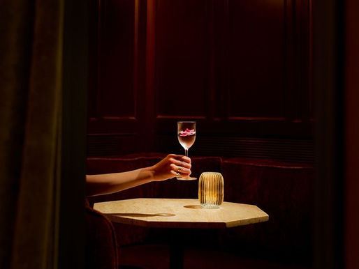 Moody, softly lit interior with a hand holding a glass at a small table, evoking restraint, elegance and intentional brand experience.