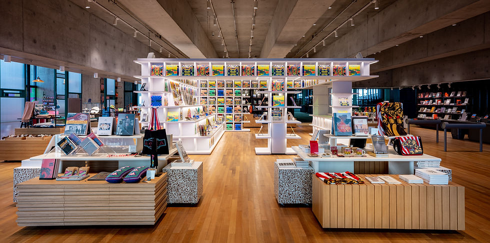 An image of a boutique shop. There are wooden floors and a concrete ceiling, as well as a series of pedestals that hold books, notebooks, bags, and other items.