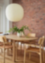 A wooden table and chairs sit in front of a brick wall. A large, circular paper lantern hangs from the ceilings and greenery sits in a vase on the table.