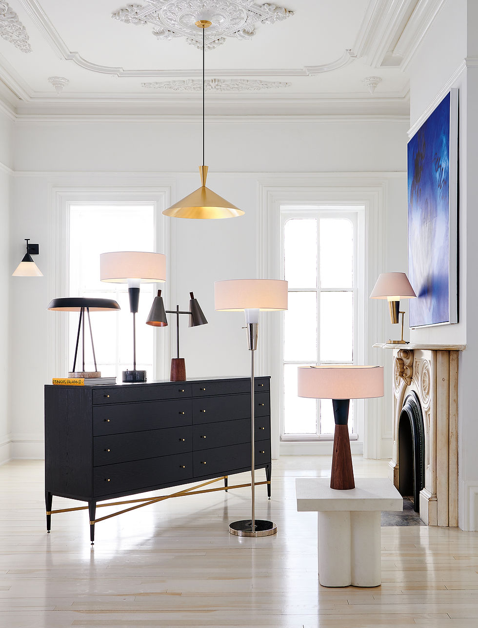 Ceiling medallion with brass pendant light hanging over midcentury table and floor lamps, dresser and bench, blue painting with hardwood floors white room with windows