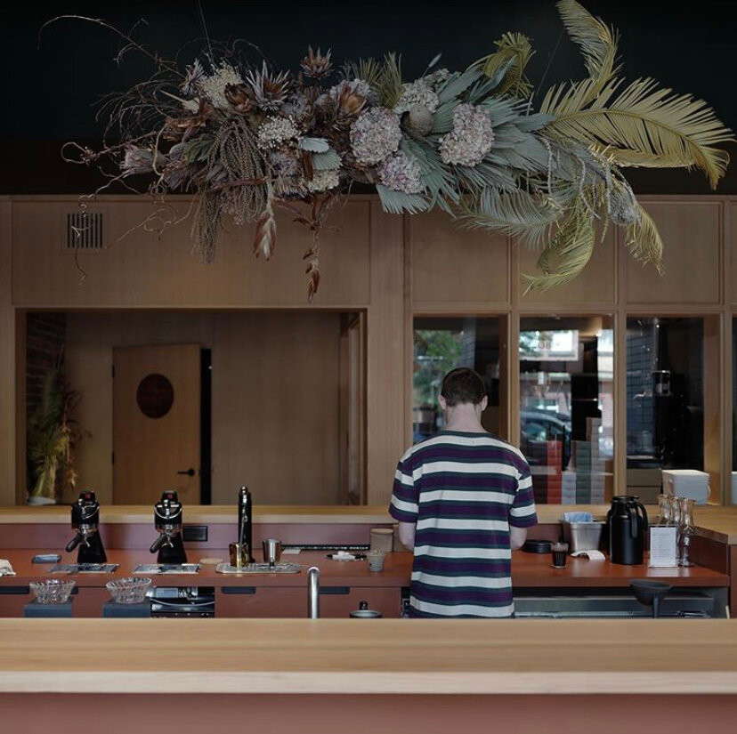 Interior of a cafe clad in light-colored wood. A man in a striped shirt stands at a coffee bar with his back to us and a large floral installation hangs overhead.