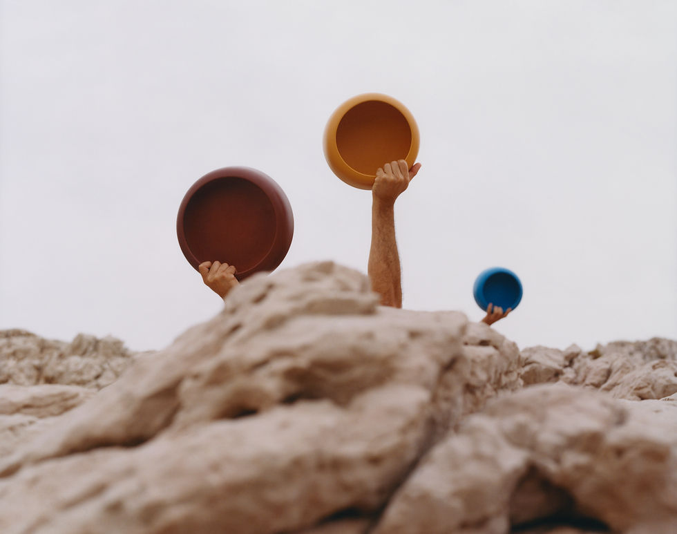 Three arms reach up from behind a rock. Each one is holding a bowl: one red, one yellow, one blue.