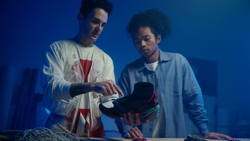 Two men stand in a blue-lit room, both looking at a black sneaker that one of them is holding.