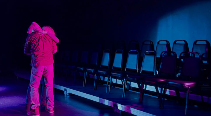 Two actors stage kiss in a pink and blue light in the background you can see the black box theatre audience.