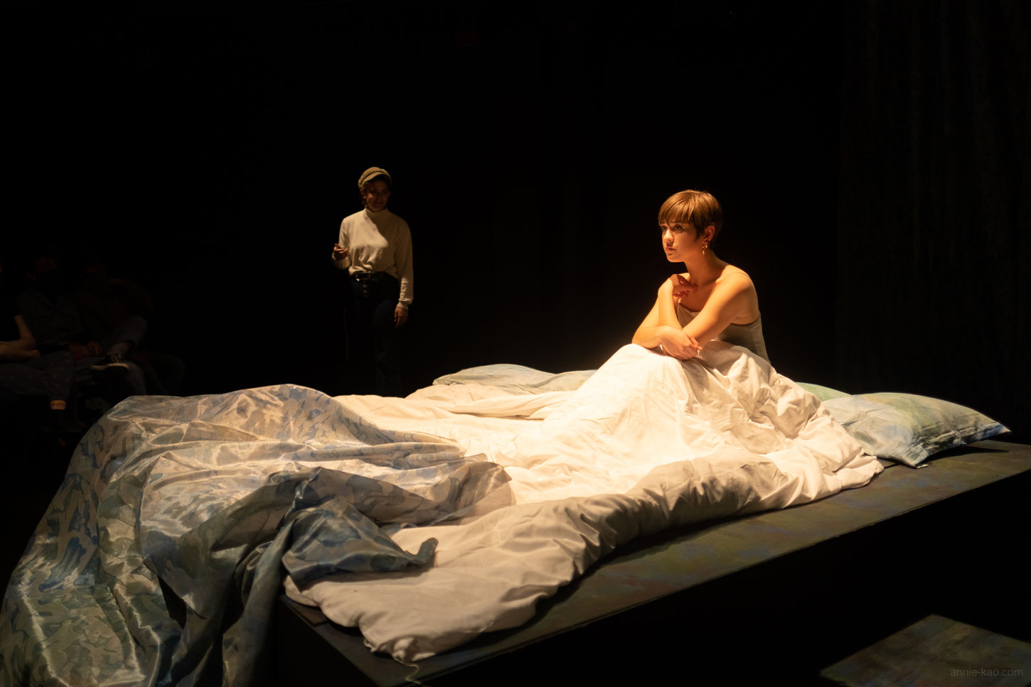 Gertrude (a white presenting woman) sits in her messy bed in a slip. She looks guilty. Her bed has a white duvet and a sheet that matches the curtains (blue and green). She is lit in bright white from above. Behind her in the distance is Horatio, in a top light, a page boy cap and an off white long sleeve shirt. The rest of the scene is black