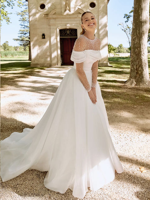 Abito da sposa ball gown in organza con scollo off-the-shoulder drappeggiato, corpetto asimmetrico, gonna ampia plissettata con strascico chapel e bordo in crine, abbinato a coprispalle in tulle con perle e maniche lunghe