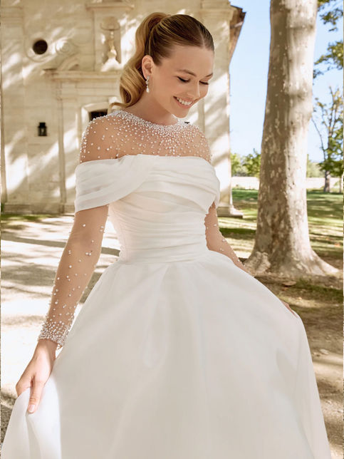 Abito da sposa ball gown in organza con scollo off-the-shoulder drappeggiato, corpetto asimmetrico, gonna ampia plissettata con strascico chapel e bordo in crine, abbinato a coprispalle in tulle con perle e maniche lunghe