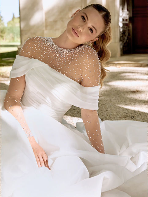 Abito da sposa ball gown in organza con scollo off-the-shoulder drappeggiato, corpetto asimmetrico, gonna ampia plissettata con strascico chapel e bordo in crine, abbinato a coprispalle in tulle con perle e maniche lunghe