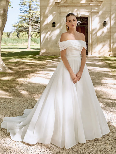 Abito da sposa ball gown in organza con scollo off-the-shoulder drappeggiato, corpetto asimmetrico, gonna ampia plissettata con strascico chapel e bordo in crine, abbinato a coprispalle in tulle con perle e maniche lunghe