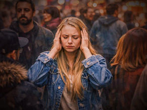 Junge Frau steht in einer belebten, unscharfen Menschenmenge und hält sich mit geschlossenen Augen die Hände an die Schläfen, als Zeichen von Reizüberflutung und sensorischem Overload in einer lauten, urbanen Umgebung.