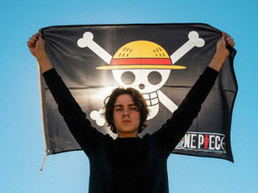 Ein junger Mensch hält eine schwarze One-Piece-Piratenflagge mit Strohhut-Totenkopf über dem Kopf, vor klarem blauem Himmel.