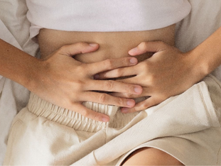 Frau liegt auf dem Bett und hält sich mit beiden Händen den Unterbauch – Symbolbild für Menstruationsbeschwerden oder Bauchschmerzen.