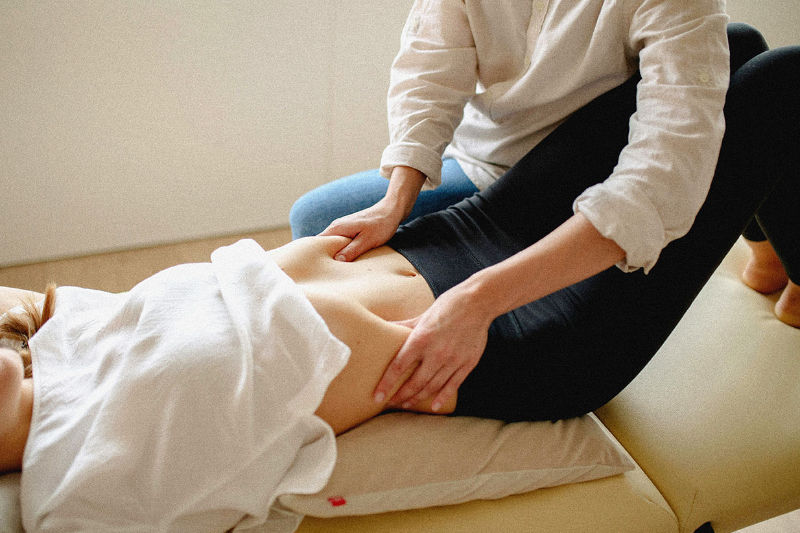 Eine Frau liegt auf einer Massageliege, während eine zweite Person mit den Händen sanft den unteren Bauchbereich berührt.