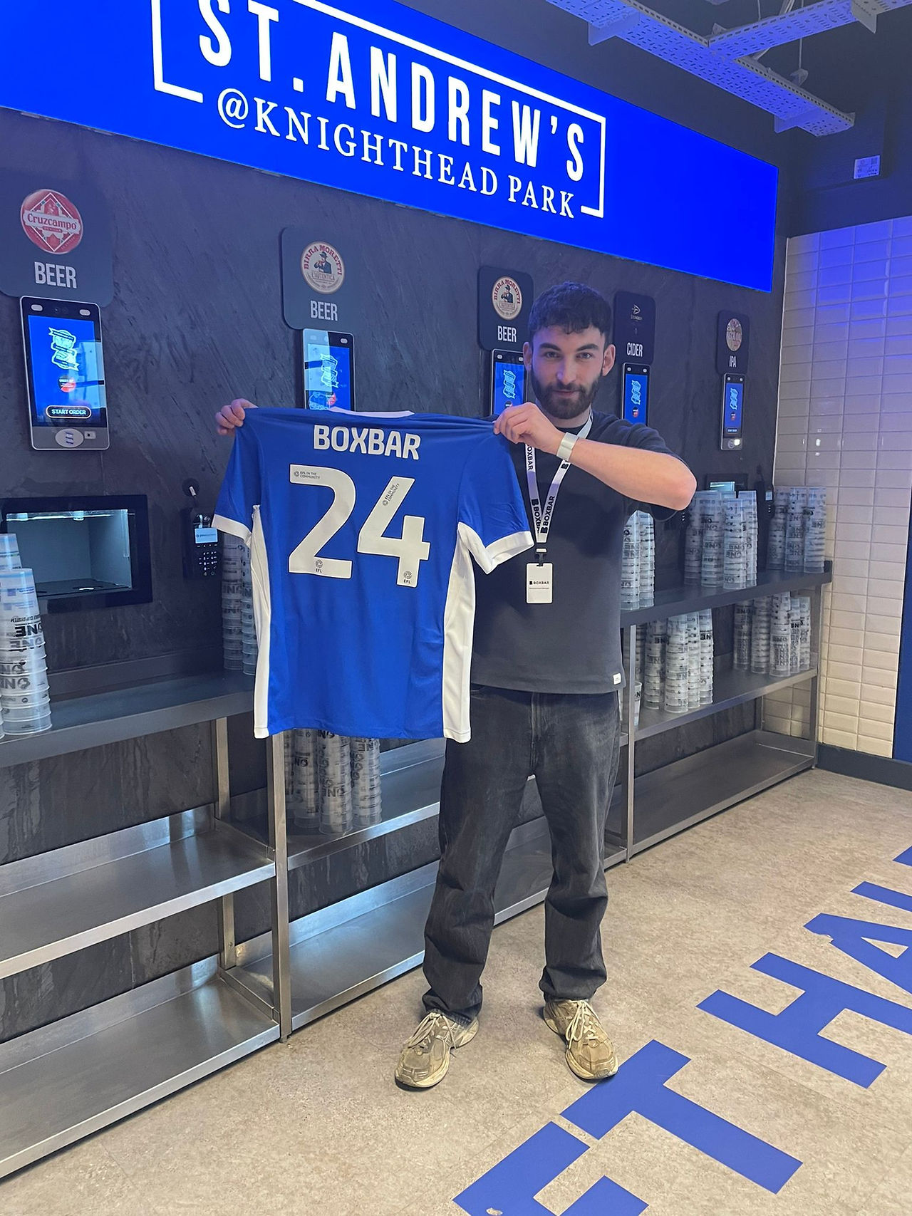 Man holds up blue football shirt with BOXBAR 24 printed on the back