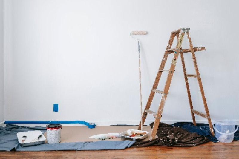 Interior painting setup with a wooden ladder, paint roller extended towards a white wall, a paint can, tray, color swatches, and protective drop cloths on a hardwood floor.
