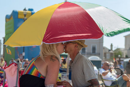 Couple kissing under rainbow umbrella at sunny outdoor Pride event.