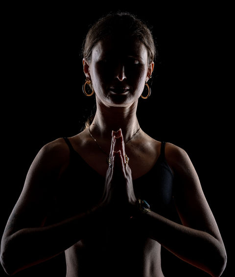 Woman in prayer pose, backlit with strong shadows on a dark background.
