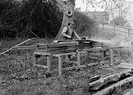 Man operating a portable sawmill, cutting logs, generating sawdust outdoors.